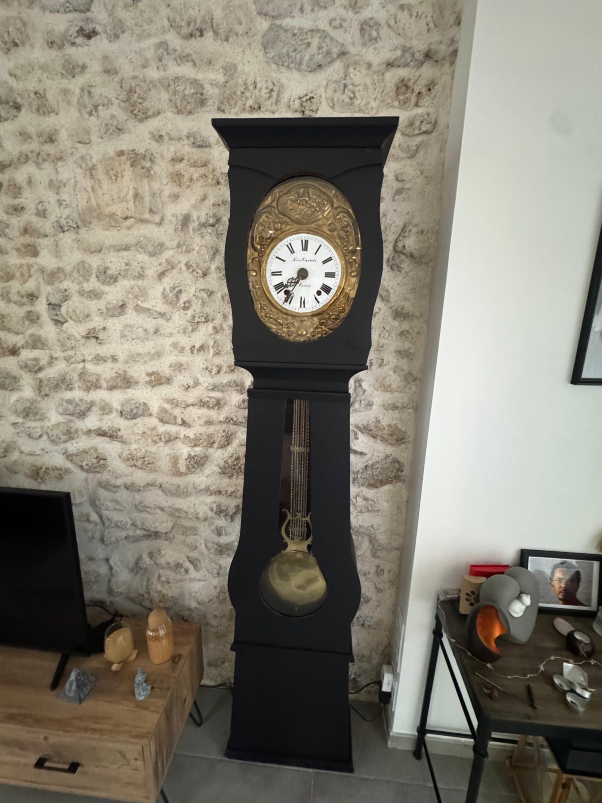 Horloge comtoise à caisse noire et lentille de laiton, devant un mur de pierre